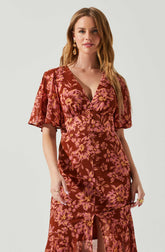 A woman is wearing a floral dress with a deep V neckline. The dress features a pattern of pink and yellow flowers on a reddish-brown background. The dress has short, loose sleeves and a fitted waist, with a slit at the front that reveals part of her legs.